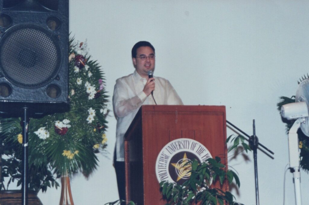 Alan Peter Cayetano speaks at a podium with the Polytechnic University of the Philippines seal, likely addressing students or faculty.