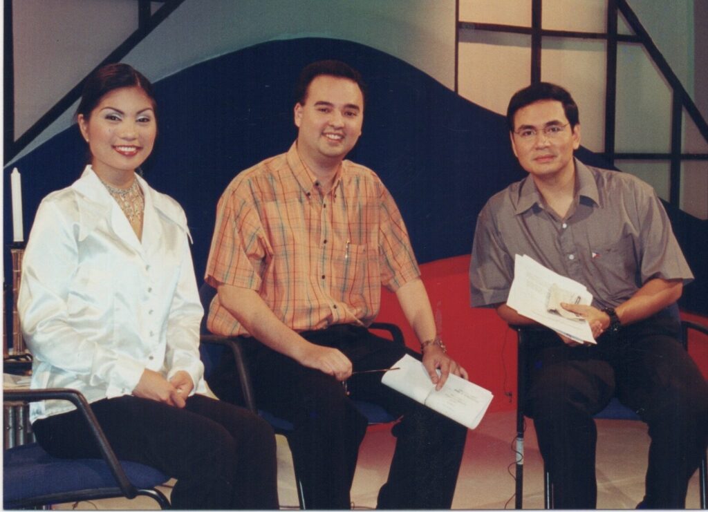 Alan Peter Cayetano is on a TV set with two other people, possibly for a show or interview, in a studio environment.