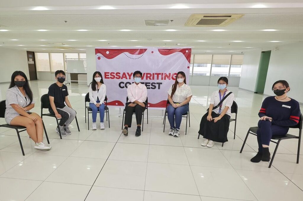 The photo shows participants seated at an essay writing contest, posing in front of the event banner.